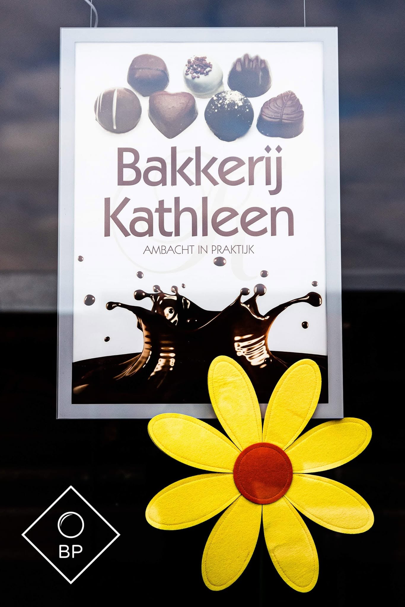 Het bord aan het winkelraam: Bakkerij Kathleen, ambacht in praktijk, met pralines en chocolade